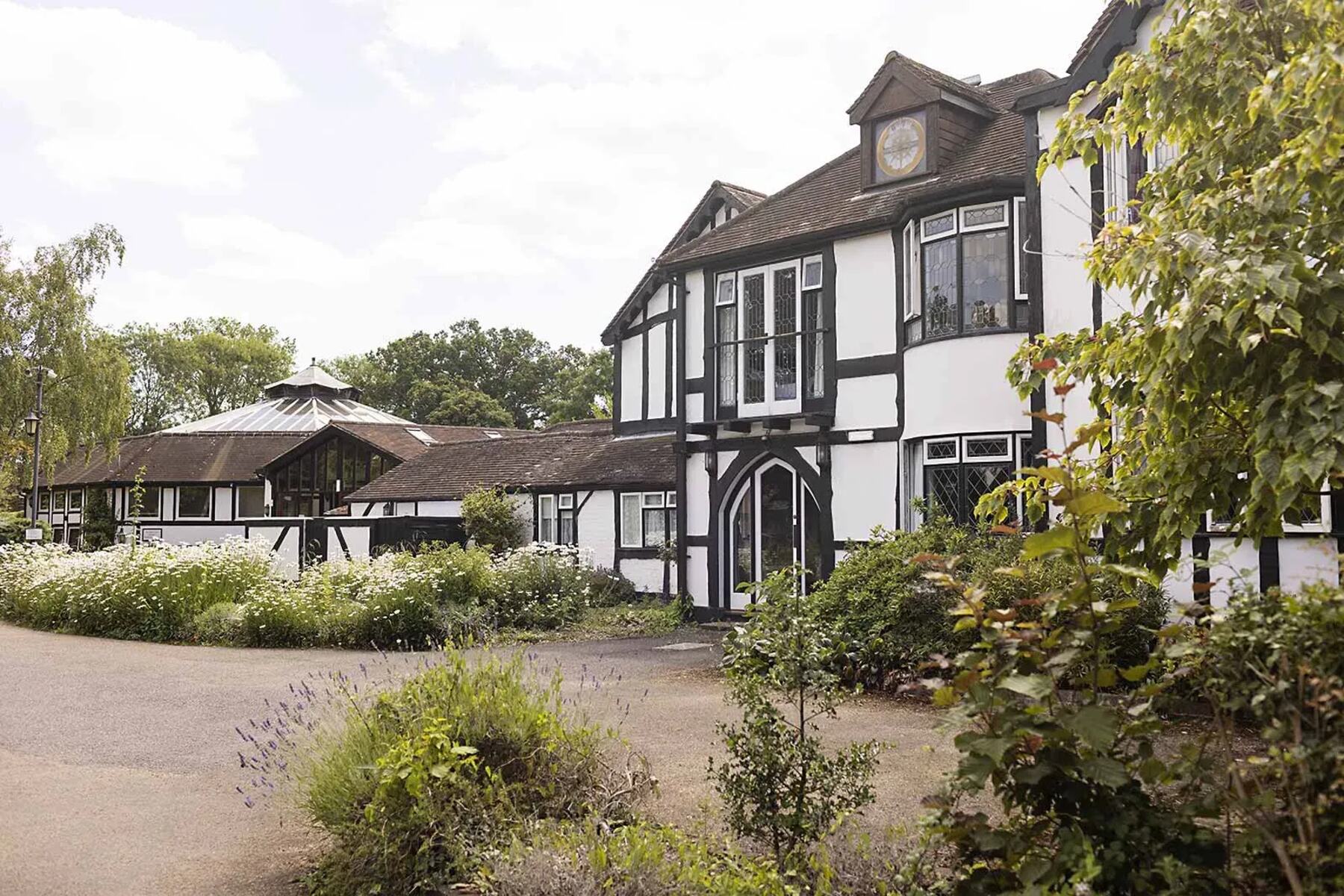 The entrance to Cardinal Clinic: a white and black timbered building with a tiled roof, approached across a gravel drive edged with wildflowers.